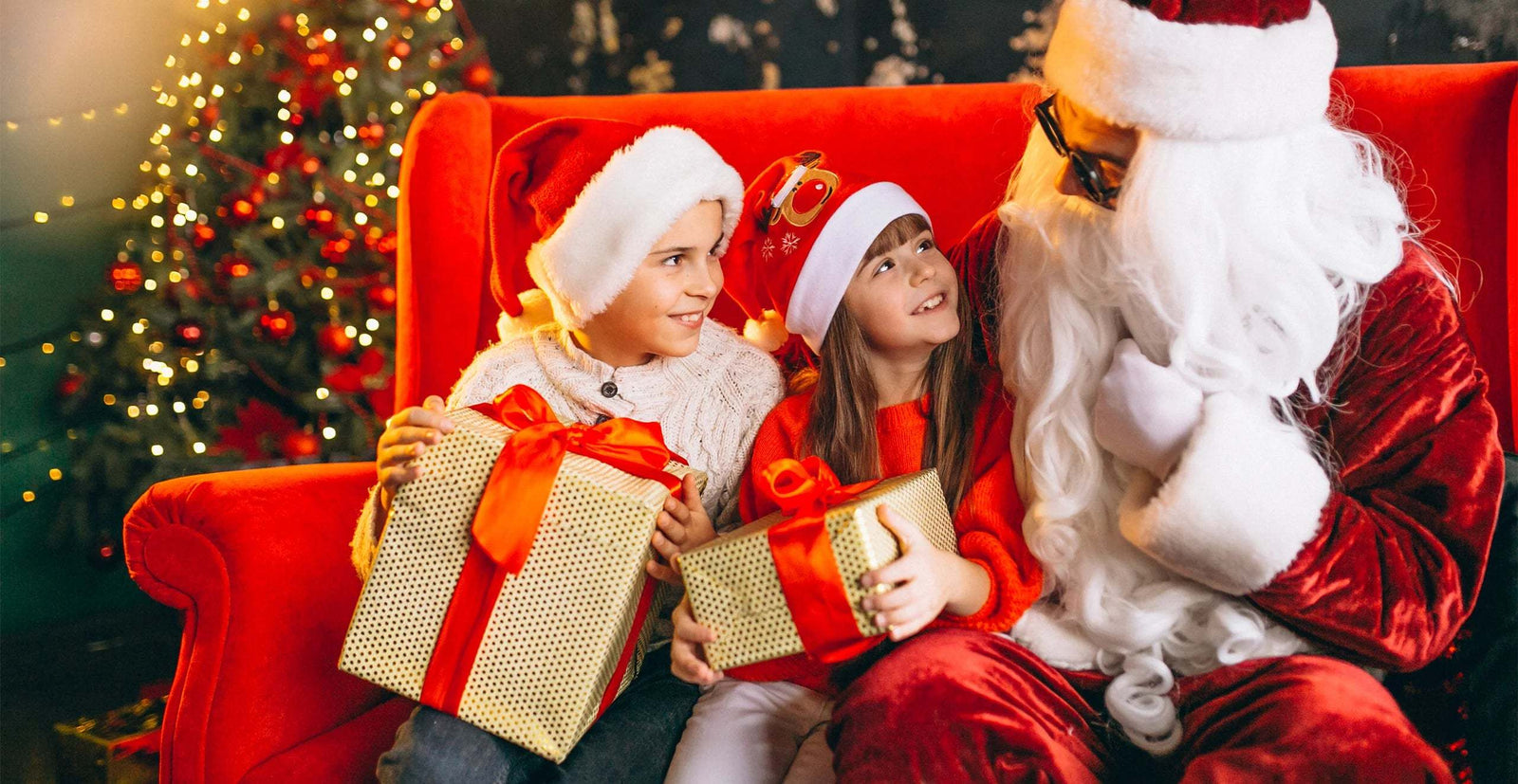 Two children in Santa hats holding Christmas gifts while sitting next to Santa Claus on a red couch under a decorated Christmas tree